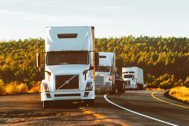 A line of white semi-trucks driving on a winding road surrounded by trees.